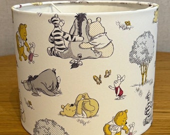 Puede incluir: Una pantalla de lámpara blanca con un patrón de Winnie the Pooh. El patrón presenta a Winnie the Pooh, Piglet, Eeyore, Tigger y otros personajes de las historias de Winnie the Pooh. La pantalla de la lámpara está hecha de tela y tiene un fondo blanco.