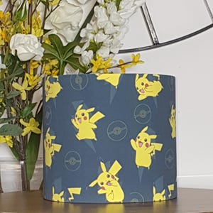 May include: A cylindrical lampshade featuring a repeating pattern of yellow Pikachu characters, Poké Balls, and lightning bolts on a dark blue background. The lampshade is a home decor item, likely for a table or floor lamp.
