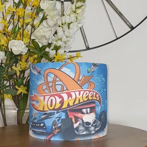 Könnte beinhalten: Zylindrischer Lampenschirm mit Hot Wheels-Logo und Grafiken von Rennwagen auf blauem Hintergrund. Das Design umfasst ein rot-oranges Logo mit einem Auto im Schädel-Design. Der Lampenschirm steht auf einer Holzoberfläche.