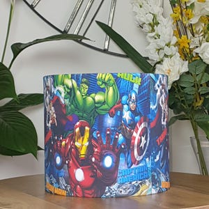 Pode incluir: Abajur cilíndrico com uma colagem de super-heróis da Marvel, incluindo Homem de Ferro, Hulk e Capitão América, contra um cenário urbano. O design incorpora cores vibrantes como vermelho, verde e azul, criando uma estética dinâmica e atraente.