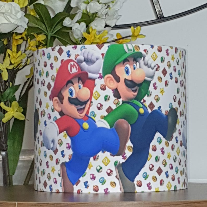 May include: Cylindrical lampshade featuring the characters Mario and Luigi from the Super Mario Bros. video game. Mario is wearing a red hat and Luigi a green hat. The background is white with colorful game-themed icons.