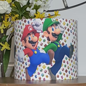 May include: Cylindrical lampshade featuring Mario and Luigi from the Super Mario Bros. franchise. The characters are depicted in their signature outfits, with a repeating pattern of game-related icons on a white background. The lampshade is a decorative item.