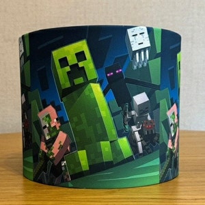 Op de afbeelding: Een lampenkap met Minecraft-thema met een groene Creeper, een zwarte Enderman, een witte geest en een roze Steve-personage. De lampenkap heeft een donkerblauwe achtergrond met groene en bruine blokken.
