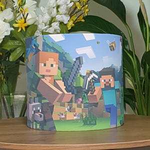 Op de afbeelding: Een cilindrische lampenkap met een Minecraft-thema. De lampenkap toont twee Minecraft-personages, een wolf, kippen en een varken. De achtergrond is een groen en bruin landschap met een blauwe lucht.