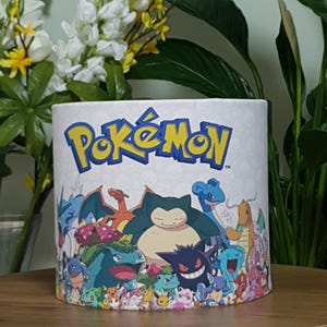Könnte beinhalten: Ein weißer Lampenschirm mit dem Pokemon-Logo und einem bunten Bild verschiedener Pokemon-Charaktere, darunter Snorlax, Glurak und Pikachu.