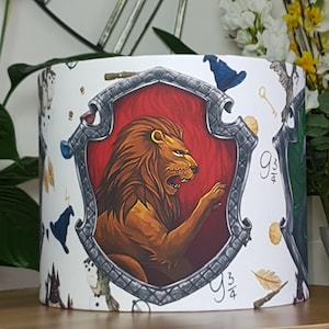 Puede incluir: Pantalla de lámpara cilíndrica blanca con una ilustración detallada de un león dentro de un escudo, que representa a Gryffindor. El diseño incluye otros símbolos de la casa, un sombrero de mago azul y el número 9 3/4.