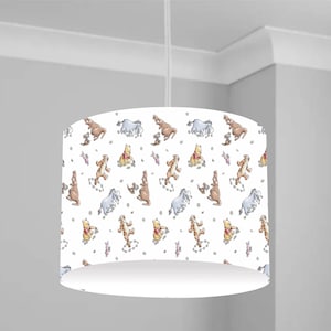 Puede incluir: Pantalla de lámpara colgante blanca con un patrón repetido de personajes de dibujos animados, incluyendo un oso amarillo, un tigre, un burro y un oso marrón. La pantalla es cilíndrica y cuelga de un cable blanco.