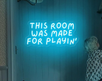 Op de afbeelding: Een blauw neonbord met de tekst "This room was made for playin'" hangt aan een muur in een kinderkamer. Een kind staat op een bankje en reikt naar een plank.