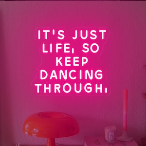 May include: Pink neon sign with the text "IT'S JUST LIFE, SO KEEP DANCING THROUGH." The sign is mounted on a pink wall with a lamp and books on a shelf below.