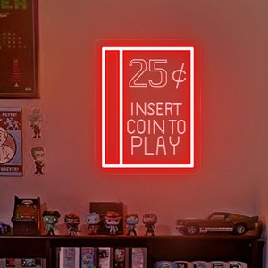 Pode incluir: Uma placa de néon vermelha com letras brancas que diz "25¢ Insira uma moeda para jogar". A placa está montada em uma parede acima de uma prateleira com figuras de videogame e cartuchos de videogame.