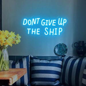 May include: A blue neon sign with the text "DON'T GIVE UP THE SHIP" hanging on a light blue wall above a striped couch with pillows.