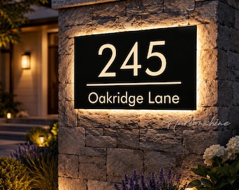 Puede incluir: Un letrero rectangular negro de número de casa montado en un pilar de piedra, con el texto blanco iluminado 245 y Oakridge Lane. El letrero está retroiluminado con un brillo cálido, resaltando la dirección sobre la superficie metálica oscura.