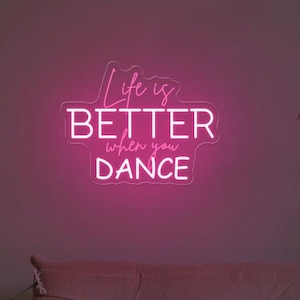 Peut inclure: Un panneau lumineux rose néon avec le texte "Life is Better when you DANCE" sur un mur rose avec un canapé au premier plan.