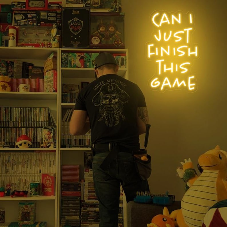 Puede incluir: Una persona lleva una camiseta negra con un gr&aacute;fico de calavera y huesos cruzados y el texto "Seize My Soul" se encuentra frente a una estanter&iacute;a llena de videojuegos. Un letrero de ne&oacute;n amarillo en la pared dice "Can I Just Finish This Game".
