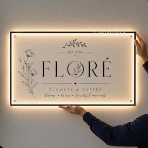 May include: Illuminated sign with the word "FLORÉ" in a stylish font, with the text "FLOWERS & COFFEE" below. The sign also includes the text "blooms • brews • beautiful moments" and "EST. 2024". The sign is black framed and held by a person.