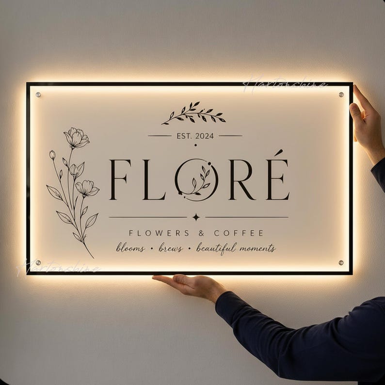 May include: A rectangular, backlit wall sign with a cream-colored face and black frame. The sign features the text "FLOR&Eacute;", "EST. 2024", "FLOWERS & COFFEE", and "blooms &bull; brews &bull; beautiful moments" alongside a black line-art floral illustration.