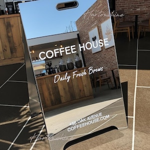 Könnte beinhalten: Ein verspiegeltes A-Rahmen-Schild für ein Café. Das Schild zeigt in weißer Schrift "COFFEE HOUSE" und "Daily Fresh Brews". Die Adresse "456 OAK AVENUE COFFEEHOUSE.COM" ist ebenfalls zu sehen. Das Schild spiegelt das Innere und Äußere des Cafés wider.