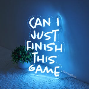 Puede incluir: Un letrero de ne&oacute;n blanco con el texto "Can I just finish this game" sobre un fondo azul.