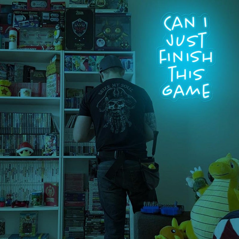 Puede incluir: Una persona se encuentra de pie frente a una pared con un letrero de ne&oacute;n que dice "Can I Just Finish This Game". La persona lleva una camiseta negra con un gr&aacute;fico de calavera y huesos cruzados y las palabras "Seize My Soul". La persona est&aacute; de pie frente a una estanter&iacute;a llena de videojuegos.