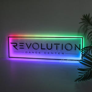 May include: A rectangular LED sign featuring the black text REVOLUTION DANCE CENTER centered on a white background. The sign is framed by a glowing border that transitions through a spectrum of colors including red, green, blue, and purple.