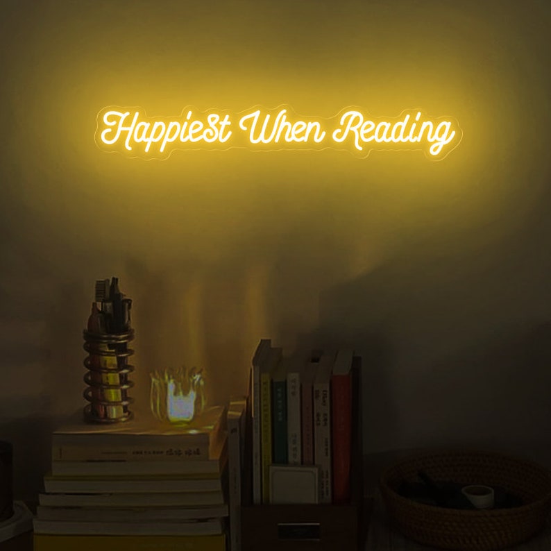 K&ouml;nnte beinhalten: Ein gelbes Neon-Schild mit dem Text "Happiest When Reading" an einer Wand mit B&uuml;cherregalen und einer Lampe.