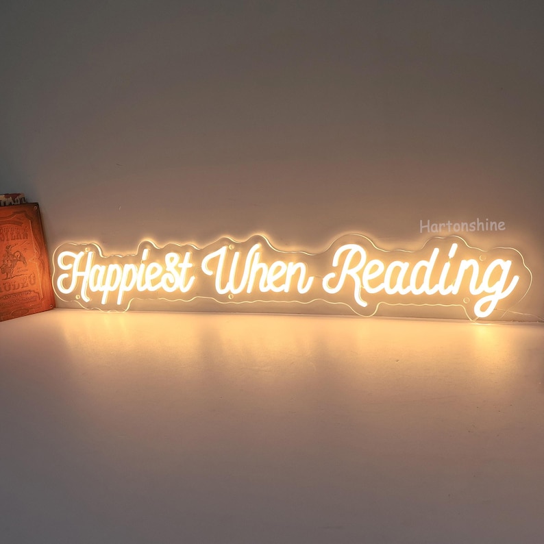 K&ouml;nnte beinhalten: Wei&szlig;es Neon-Schild mit dem Text "Happiest When Reading" in einer kursivschriftlichen Schriftart.