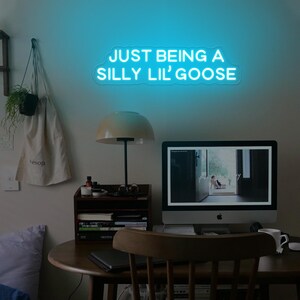 May include: A blue neon sign with the text "JUST BEING A SILLY LIL' GOOSE" hanging on a wall above a desk with a lamp, a computer, and a chair.