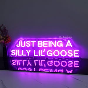 May include: Purple neon sign with the text "JUST BEING A SILLY LIL' GOOSE" in white. The sign is illuminated and has a cloudy outline.