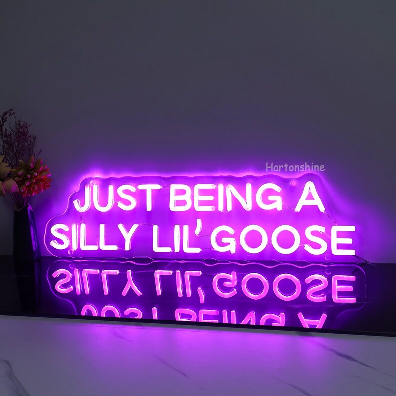 May include: Purple neon sign with the text "JUST BEING A SILLY LIL' GOOSE" in white. The sign is illuminated and has a cloudy outline.