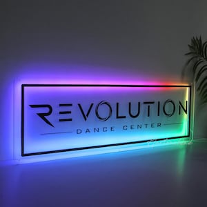 May include: A rectangular acrylic sign featuring the black text REVOLUTION DANCE CENTER. The sign is illuminated with a vibrant, multi-colored LED light border that transitions through shades of purple, blue, and green along the edges.