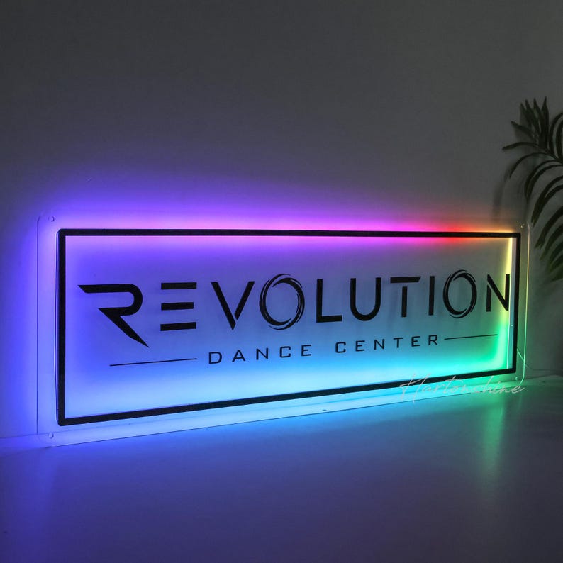 May include: A rectangular acrylic sign featuring the black text REVOLUTION DANCE CENTER. The sign is illuminated with a vibrant, multi-colored LED light border that transitions through shades of purple, blue, and green along the edges.