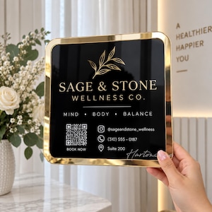 以下が含まれることがあります： 金色の金属フレームが付いた正方形の黒い看板。SAGE & STONE WELLNESS CO.の文字、MIND • BODY • BALANCEという言葉、QRコード、Instagramのハンドルネーム、電話番号、スイート番号が金色の文字とグラフィックで表示されています。