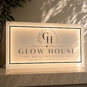 Peut inclure: Panneau lumineux pour "Glow House Aesthetics". Le panneau rectangulaire présente un cadre noir et un fond crème. Le texte comprend le nom de l'entreprise, un logo et les mots "Enhance, Renew, Glow".