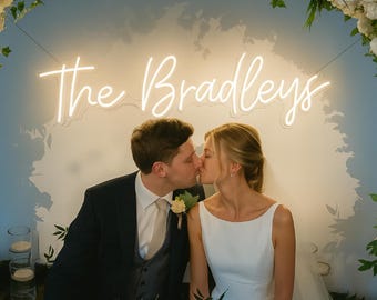 May include: A newly married couple kissing beneath a neon sign that reads "The Bradleys". The man is in a suit, and the woman wears a white wedding dress. The scene is decorated with flowers, creating a romantic atmosphere.