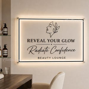 May include: A rectangular wall sign with a cream-colored background and black lettering that reads REVEAL YOUR GLOW, Radiate Confidence, and BEAUTY LOUNGE. The sign features a black line drawing of a profile and is illuminated by backlighting.