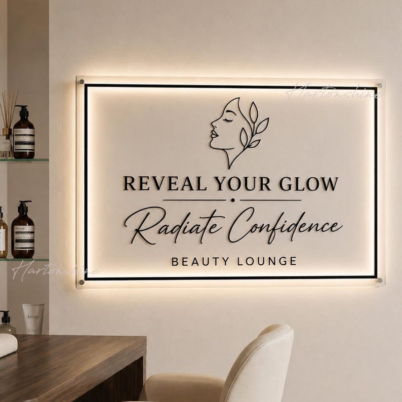 May include: A rectangular wall sign with a cream-colored background and black lettering that reads REVEAL YOUR GLOW, Radiate Confidence, and BEAUTY LOUNGE. The sign features a black line drawing of a profile and is illuminated by backlighting.