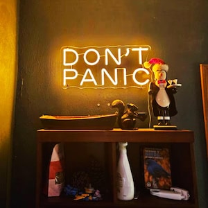 Könnte beinhalten: Ein gelbes Neon-Schild mit den Worten "DON'T PANIC" an einer dunkelbraunen Wand. Ein Holzregal mit einem Holzboot, einer Eichhörnchenfigur, einer Flüssigkeitsflasche, einem Buch über Vögel und einer Pinguinfigur.