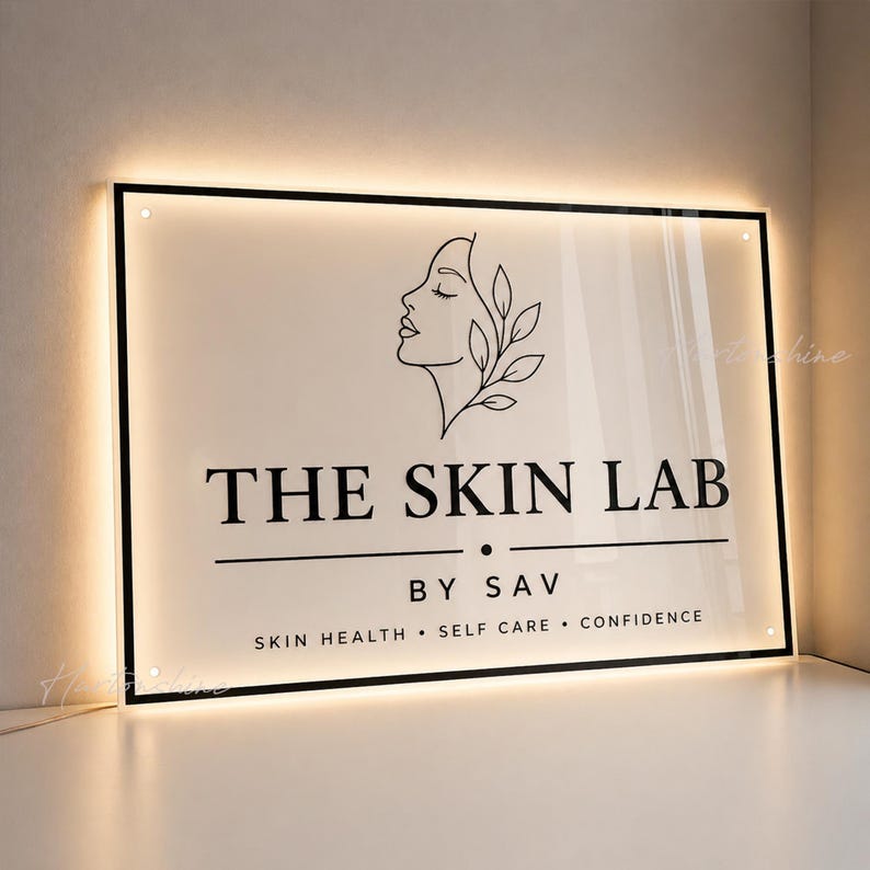 May include: A rectangular, backlit acrylic sign featuring a black line-art illustration of a profile face with leaves. The text reads THE SKIN LAB BY SAV, followed by the words SKIN HEALTH, SELF CARE, and CONFIDENCE, all in a black serif font.