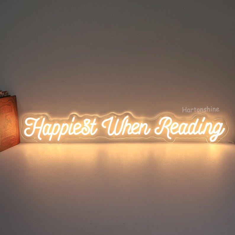 K&ouml;nnte beinhalten: Ein wei&szlig;es Neon-Schild mit dem Text "Happiest When Reading" auf wei&szlig;em Hintergrund.