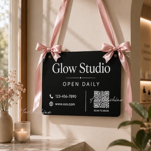 Pode incluir: Placa retangular preta com o texto "Glow Studio" em branco, pendurada por fitas rosa amarradas em laços. A placa inclui informações de contato e um código QR para reservas.