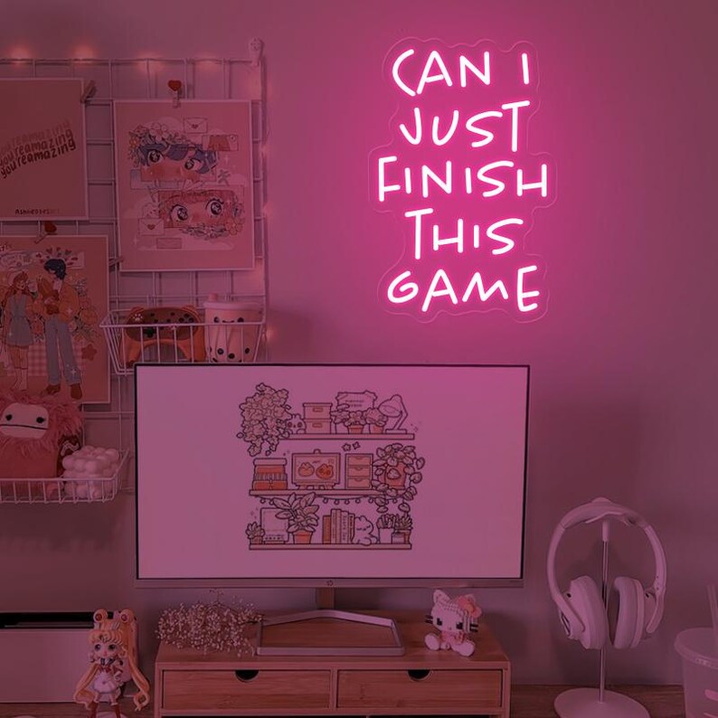 Puede incluir: Un letrero de ne&oacute;n rosa con el texto "CAN I JUST FINISH THIS GAME" en una pared rosa. El letrero est&aacute; sobre un monitor de computadora con una imagen de dibujos animados de un estante con plantas y libros. Hay un auricular blanco en un soporte a la derecha del monitor.