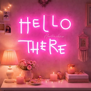 Puede incluir: Un letrero de neón rosa brillante que dice "HELLO THERE" en una fuente cursiva. El letrero está montado en una pared rosa, con una lámpara, flores, velas y otros elementos decorativos en una superficie blanca debajo.