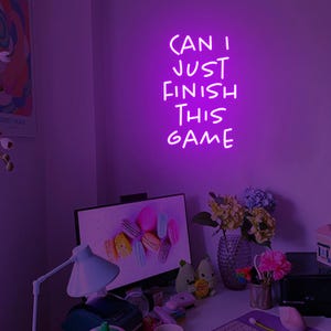 Puede incluir: Un letrero de ne&oacute;n morado con el texto "CAN I JUST FINISH THIS GAME" en una pared morada en una habitaci&oacute;n con un escritorio y una computadora.