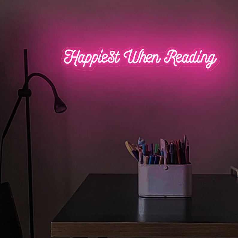 K&ouml;nnte beinhalten: Rosa Neon-Schild mit dem Text "Happiest When Reading" an einer rosa Wand. Im Vordergrund ein Schreibtisch mit einer Lampe und einem wei&szlig;en Beh&auml;lter gef&uuml;llt mit bunten Stiften.