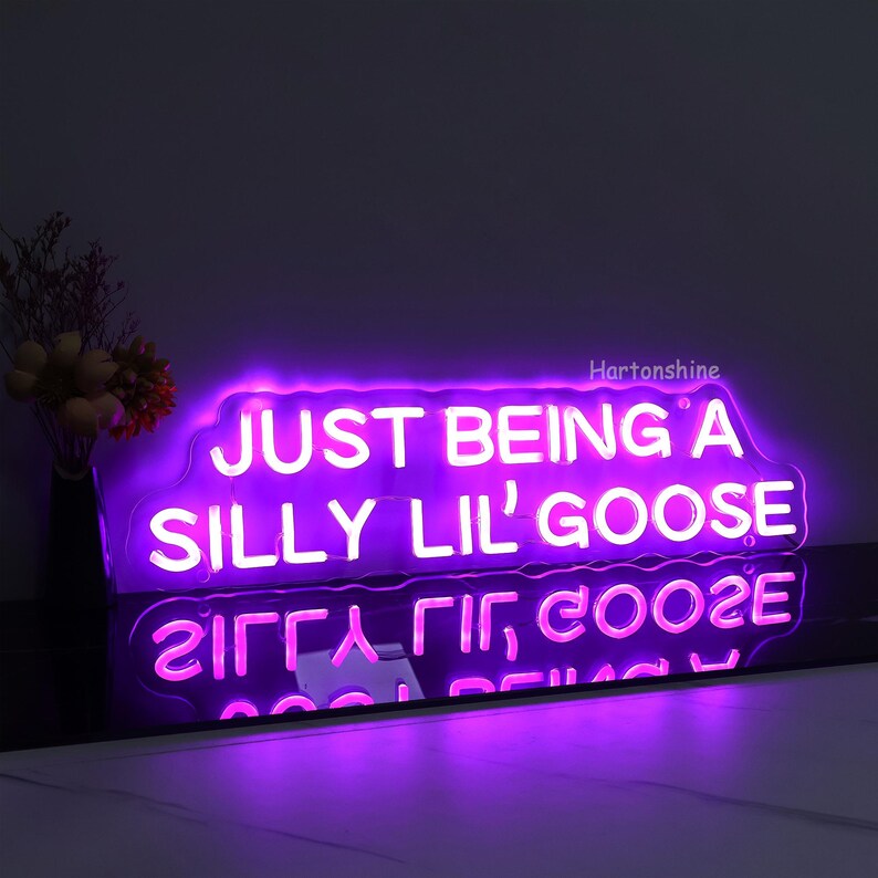 May include: Purple neon sign with a white outline that reads "JUST BEING A SILLY LIL' GOOSE". The sign is on a black surface and is reflected in the surface.