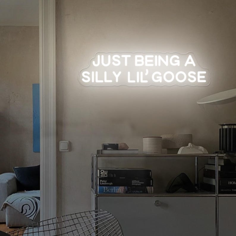 May include: White neon sign with the text "JUST BEING A SILLY LIL' GOOSE" on a white wall in a room with a bookshelf and a chair.
