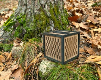 May include: A wooden cube with a checkerboard pattern on its sides. The cube has a dark frame and is set against a natural backdrop of leaves, grass, and a tree trunk.
