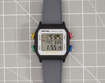 May include: A digital wristwatch with a grey textured silicone strap and a black rectangular case. The face features a world map graphic, the text RETRO WORLD TIME, and WATER RESIST. It has four colorful buttons: yellow, red, green, and blue.