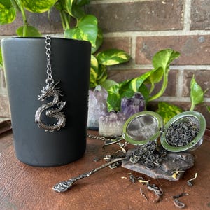 May include: A black ceramic mug with a silver dragon tea infuser hanging from the rim. A stainless steel tea strainer is open, filled with loose tea leaves. A decorative tea spoon rests nearby. The background includes plants and crystals.
