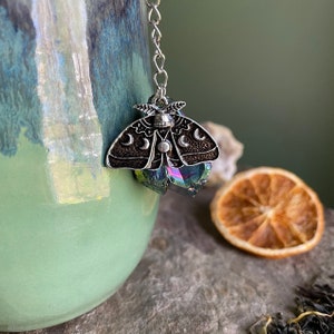 May include: Silver tea infuser with a black moth design and iridescent crystal accents. The moth has a crescent moon pattern on its wings.